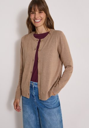 Femme souriante portant un cardigan beige boutonné sur un haut bordeaux et un jean bleu en denim, debout avec une main dans la poche.