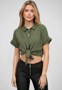 Cămașă cu mâneci scurte, cu guler, de culoare verde măslin, cu un design texturat și ribat, având un detaliu de legare în față. Asortată cu pantaloni negri cu talie înaltă.