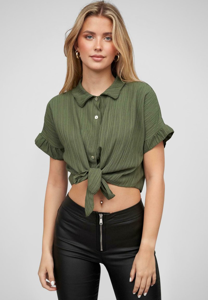 Cămașă cu mâneci scurte, cu guler, de culoare verde măslin, cu un design texturat și ribat, având un detaliu de legare în față. Asortată cu pantaloni negri cu talie înaltă.