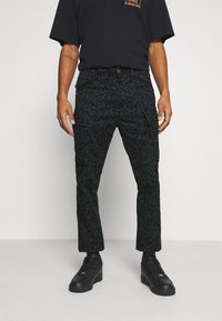 Cargo-byxor i mörkt tyg med ett subtilt blommönster. Har sidofickor och en skräddarsydd passform, ihop med svarta skor och en svart t-shirt.
