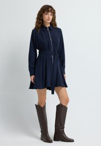 Granatowa sukienka z kołnierzykiem, długimi rękawami i zamkiem. W zestawie znajduje się dopasowany pasek oraz rozkloszowana spódnica. Zestawiona z brązowymi botkami do kolan.