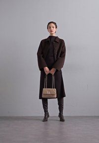 Palton maro din lână cu mâneci supradimensionate, rochie neagră ajustată, cizme din piele până la genunchi și o geantă bej matlasată cu mânere din lanț.