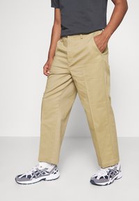 Pantalons cargo beiges avec une coupe décontractée, dotés de poches latérales et d'une texture de tissu lisse, associés à des chaussures de sport grises et blanches.