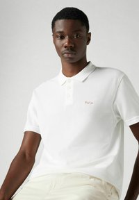 Vit polo skjorta tillverkad av texturerad bomull med krage och treknappars placket. Har en röd broderad "Paris" på vänster bröst.