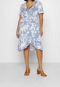 Vestido envolvente floral en azul claro con patrones blancos, mangas cortas y dobladillo con volantes; combinado con sandalias de tacón cuadrado marrones.