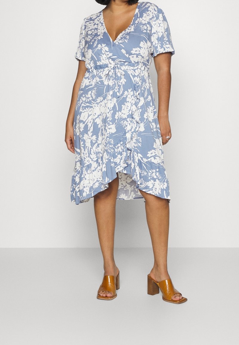 Vestido envolvente floral en azul claro con patrones blancos, mangas cortas y dobladillo con volantes; combinado con sandalias de tacón cuadrado marrones.
