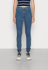 Mujer vistiendo jeans ajustados de cintura alta en azul, zapatillas negras y blancas, y una camiseta cortada en navy con rayas blancas, de pie sobre un suelo gris.