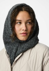 Młoda kobieta w ciemnoszarym dzianym kapturze i jasnobeżowej pikowanej kurtce, spoglądająca nieco na bok na białym tle.