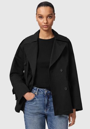 Młoda kobieta o ciemnych włosach zaczesanych do tyłu, ubrana w czarny dwurzędowy płaszcz, czarną bluzkę i niebieskie dżinsy, pozująca z jedną ręką w kieszeni.