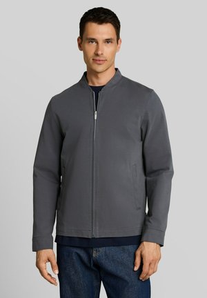 Man draagt een grijze jas met rits over een donker shirt en blauwe jeans, staand tegen een effen lichtgrijze achtergrond.