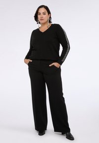 Czarny sweter v-neck z długimi rękawami, ozdobiony białym wzorem w zygzak na rękawach, w połączeniu z szerokimi czarnymi spodniami i czarnym obuwiem.