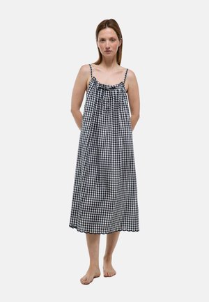 Femme debout pieds nus portant une robe midi sans manches à carreaux noirs et blancs avec fines bretelles et ourlet festonné.
