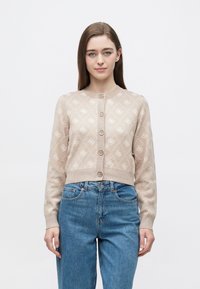 Μπεζ πλεκτή ζακέτα με διαμαντένιο μοτίβο, στρογγυλή λαιμόκοψη, εμπρός με κουμπιά και μακριά μανίκια. Συνδυασμένη με μπλε τζιν ψηλόμεσα παντελόνια.