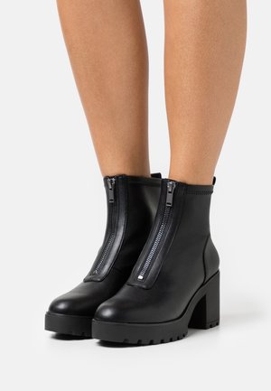 Botas de tobillo de cuero negro con dos cremalleras plateadas, un tacón grueso y una suela de goma. Textura suave con costuras reforzadas en los bordes.