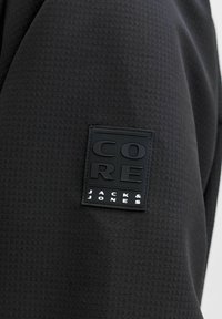 Tissu noir texturé avec un patch rectangulaire portant l'inscription « CORE » au-dessus de « JACK & JONES » en lettres blanches et noires.