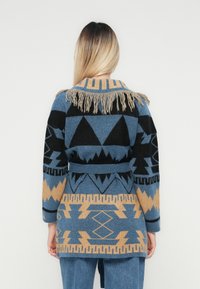 Persoană cu părul lung, purtând un cardigan tricotat cu model albastru, negru și bej, cu franjuri la guler și curea în talie, întorcându-se.