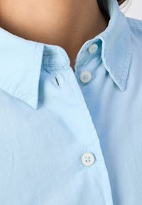 Col de chemise bleu clair boutonné, porté ouvert, montrant la peau du cou sans bijoux ni accessoires.