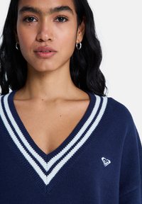 Μπλε ναυτικό πλεκτό πουλόβερ με V-λαιμόκοψη, διακοσμημένο με λευκές ρίγες και μικρό λογότυπο καρδιάς στην αριστερή πλευρά. Μαλακή υφή και άνετη εφαρμογή.