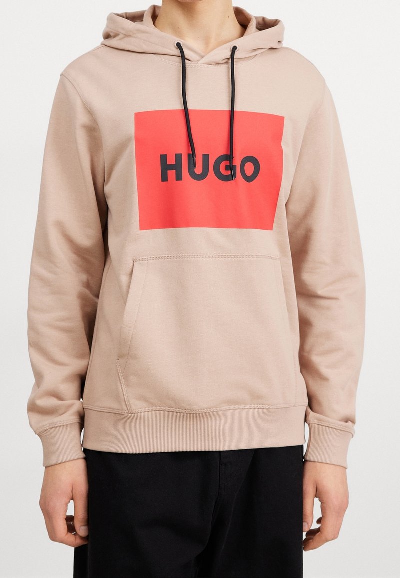 Sudadera beige de tejido suave, con un gran gráfico rectangular rojo que presenta la palabra "HUGO" en letras negras en negrita. Capucha con cordones, bolsillo frontal.