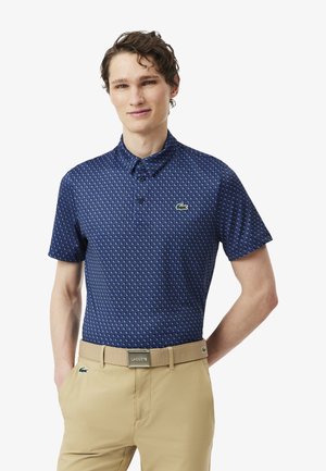 Jonge man die een blauw poloshirt met patroon en korte mouwen draagt, beige broek met een beige Lacoste-riem, staand met één hand in de zak.