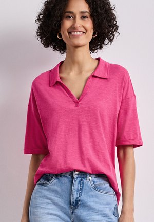Femme aux cheveux bouclés portant des boucles d’oreilles créoles dorées, une chemise rose vif à manches courtes avec col rentrée dans un jean bleu clair, souriante.