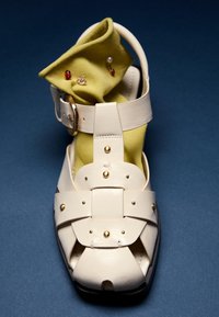 Sandale en cuir crème avec des clous dorés sur une chaussette verte pliée ornée de petites épingles coccinelle et perle, posée sur une surface bleu foncé.
