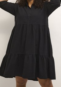 Schwarzes Hemdkleid aus Baumwolle mit einem gestuften, gerafften Saum. Verfügt über lange Ärmel und eine lockere Passform. Glatte Textur.