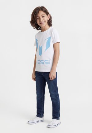 Smiling boy with curly brown hair wearing a white Messi graphic t-shirt, blue jeans, and white sneakers, standing against a white background.