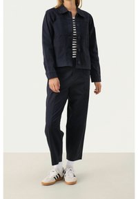 Granatowa kurtka dżinsowa z przednimi kieszeniami i zapięciem na guziki, zestawiona z dopasowanymi granatowymi spodniami oraz białymi trampkami z czarnymi paskami.