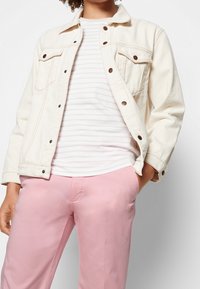 Krämfärgad denimjacka med framfickor, bruna knappar, bärs över en vit och ljusgrå randig skjorta, tillsammans med rosa byxor.