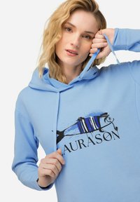 Světle modrá mikina s kapucí vyrobená z měkké tkaniny, která má grafický potisk ryby s černými a modrými pruhy a nápisem "LAURASON."