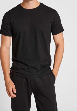 Mann trägt schlichtes schwarzes T-Shirt mit kurzen Ärmeln und schwarze Hose, steht mit einer Hand in der Tasche vor hellem Hintergrund.