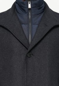 Manteau en laine gris foncé avec un col montant porté sur une veste matelassée bleu marine zippée jusqu'au cou.
