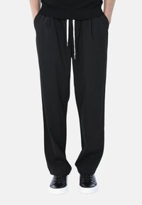 Pantaloni neri a gamba larga realizzati in tessuto morbido, con vita elastica e un cappio bianco, dotati di tasche laterali. Abbinati a sneakers nere.