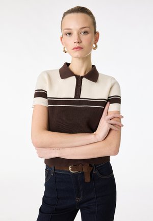 Kobieta o jasnej karnacji i włosach związanych do tyłu, ubrana w brązowo-kremową, w paski, krótkim rękawem polo oraz ciemne jeansy z brązowym paskiem.