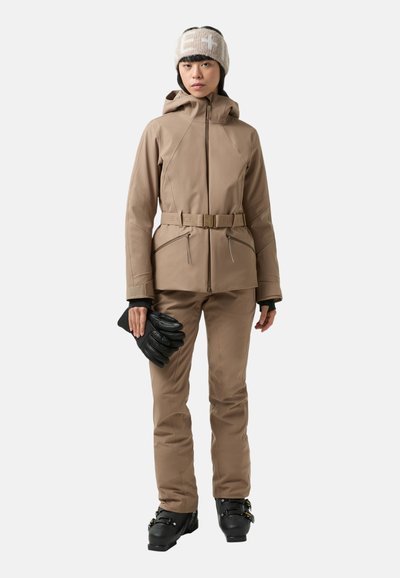 Skianzug in Beige, mit Gürtel in der Taille, Kapuze und zwei vorderen Reißverschlusstaschen. Das Model hält schwarze Handschuhe und trägt ein leichtes Strickstirnband.