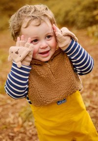 Gilet in pile marrone con zip sopra una maglietta a maniche lunghe a righe blu, pantaloni impermeabili gialli e guanti senza dita color beige con dettagli a orso.