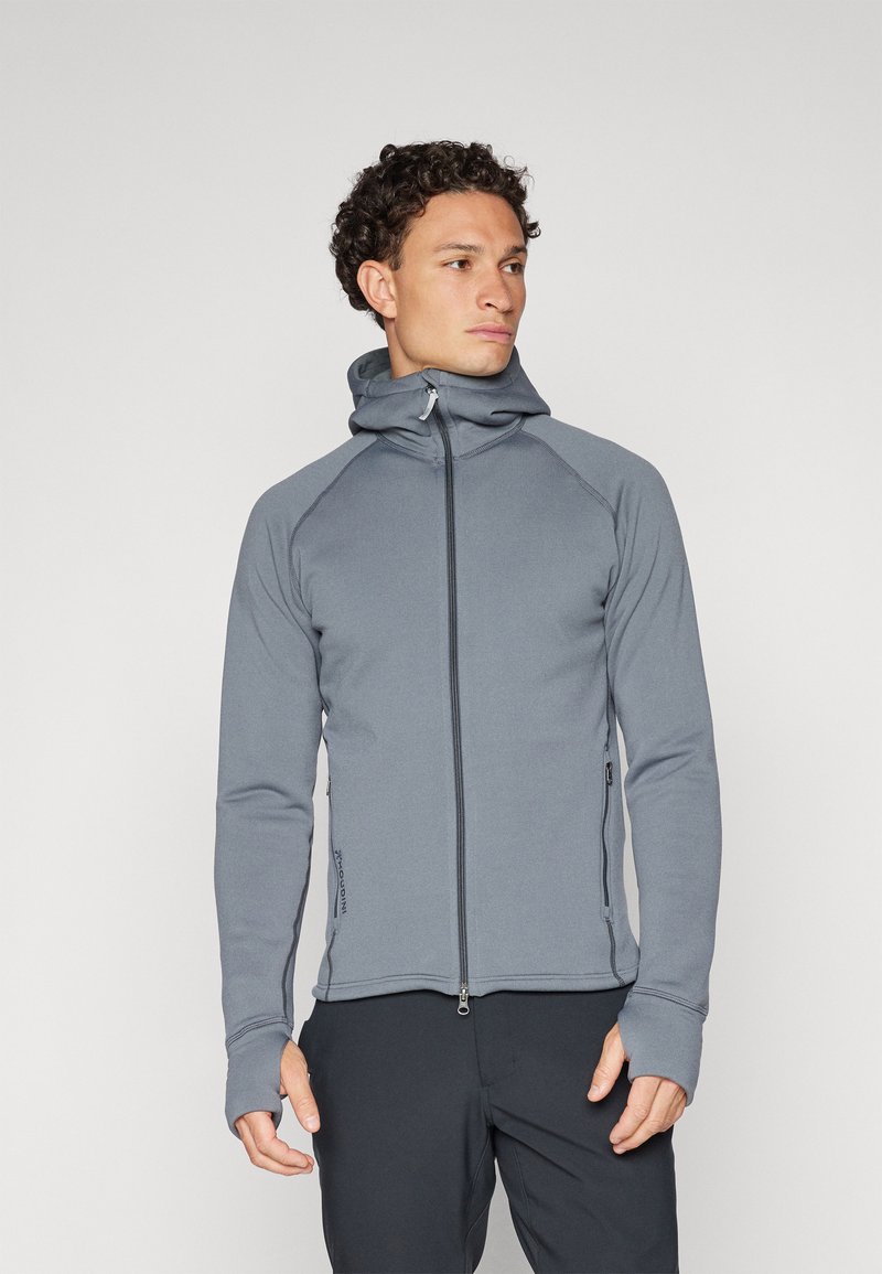 Sudadera gris con cremallera y alto cuello, mangas raglán y agujeros para los pulgares. Está hecha de una tela suave y elástica. Cuenta con una cremallera frontal y bolsillos laterales.