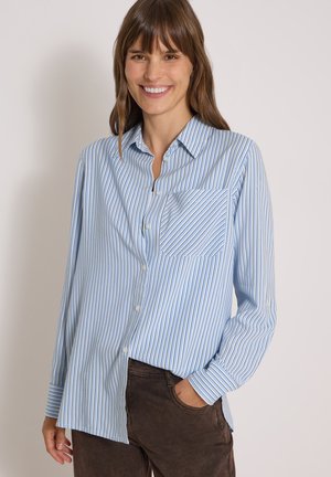Femme aux cheveux bruns souriante, portant une chemise bleu clair à manches longues à fines rayures verticales et un pantalon marron foncé, main dans la poche.