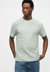 Άνδρας με σκουρόχρωμο δέρμα που φοράει ανοιχτό πράσινο κοντομάνικο υφασμάτινο καρό μπλουζάκι και μπλε τζιν, στέκεται με τα χέρια στις τσέπες.