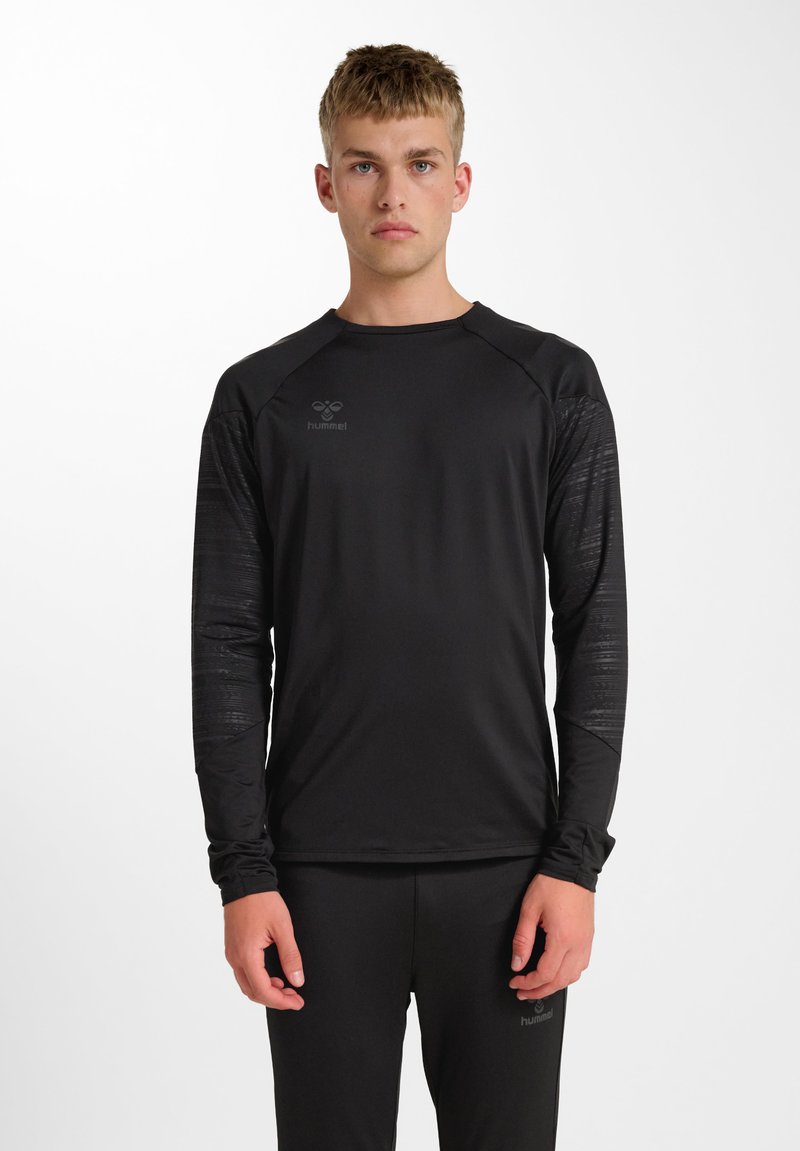 Junger Mann trägt ein schwarzes Sportshirt mit langen Ärmeln und passenden Hosen mit dezent strukturierten Ärmeln und Logo auf Brust und Oberschenkel.