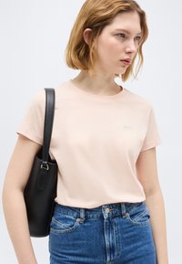 Γυναίκα που φοράει ανοιχτό ροζ μπλουζάκι με κοντά μανίκια και μπλε τζιν, κρατώντας μαύρη τσάντα ώμου, κοιτάζοντας προς τα πλάγια μπροστά σε απλό φόντο.