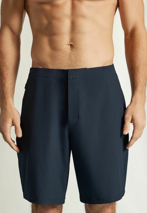 Mandlig torso iført ensfarvede mørkeblå knælange shorts med lynlåslomme på højre side, stående mod en lys baggrund.