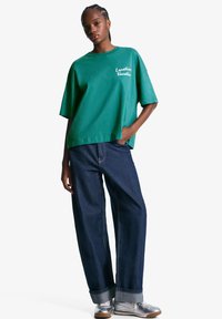 Grøn oversized t-shirt med teksten "Location: Vacation", parret med løse mørk denim jeans og sølv sneakers. Casual stil med afslappet pasform.