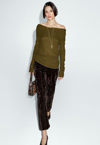 Maglione oliva off-the-shoulder, abbinato a pantaloni a gamba larga in velluto marrone scuro. Accentuato con una collana a più strati e una borsa stampata.
