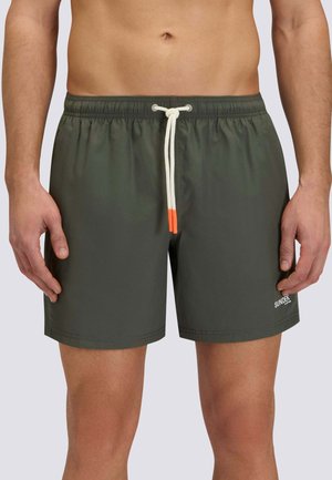 Uomo che indossa pantaloncini da bagno verde scuro con coulisse bianca con punte arancioni, in piedi davanti a uno sfondo chiaro e uniforme.