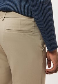 Világosbarna nadrág sima textúrával és hátsó zsebbel. A modell kék bordázott hosszú ujjú inget visel, amelynek egy részét láthatóvá teszi az ujja.