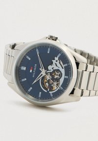 Αυτόματη ασημένια ρολόι χεριού με μπλε καντράν, ρωμαϊκούς αριθμούς, ορατά γρανάζια και μεταλλικό λουρί από την μάρκα Hilfiger.