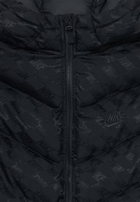 Črna Nike jakna z diagonalo embosiranih Nike logotipov, osrednji črn zadrga in prišiti Nike swoosh logotip na prsih.