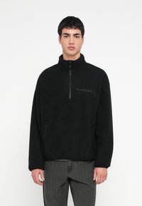 Νέος άνδρας με κοντά μαύρα μαλλιά, φοράει μαύρο φλις πουλόβερ με φερμουάρ και σκούρα γκρι τζιν, στέκεται μπροστά σε ένα απλό λευκό φόντο.